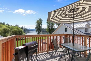 Une terrasse avec un barbecue, une table et un parasol, donnant sur un lac.