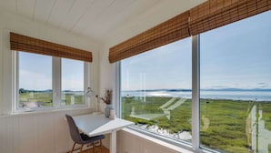 Ein Zimmer mit weißem Schreibtisch, zwei Jalousiefenstern, einem Stuhl und Blick auf ein Grasfeld sowie Wasser.
