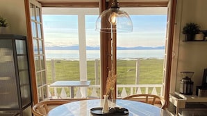 Ein runder Tisch mit sechs Stühlen, eine hängende Leuchte und Blick auf ein Gewässer durch große Fenster.
