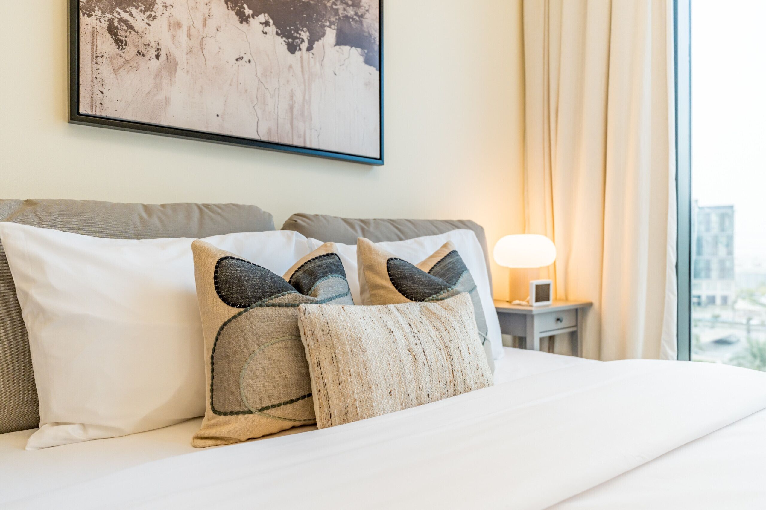 A neatly made bed with white linens and decorative pillows, a bedside table with a lamp, and a framed artwork on the wall.