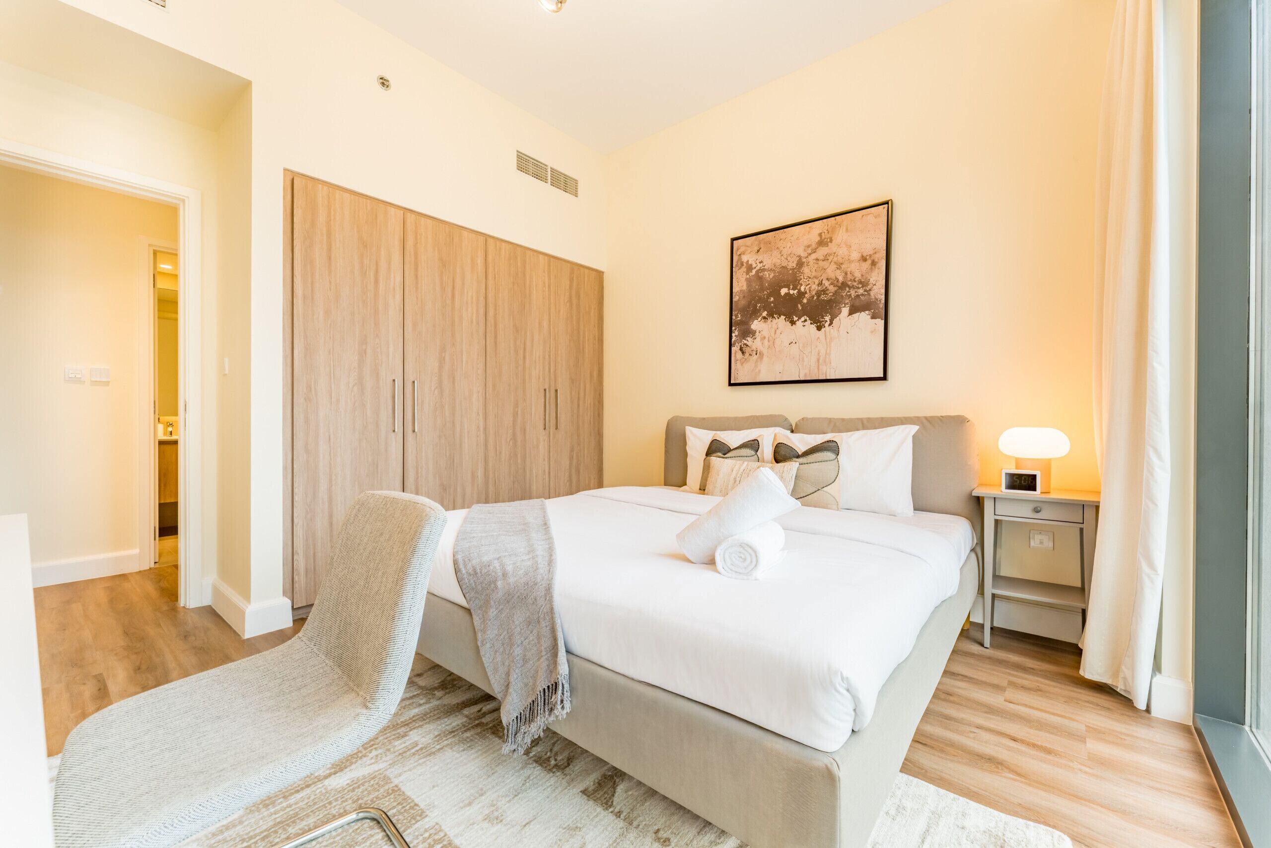 A bedroom with a bed, a chair, a wardrobe, a bedside table with a lamp, and a painting on the wall.