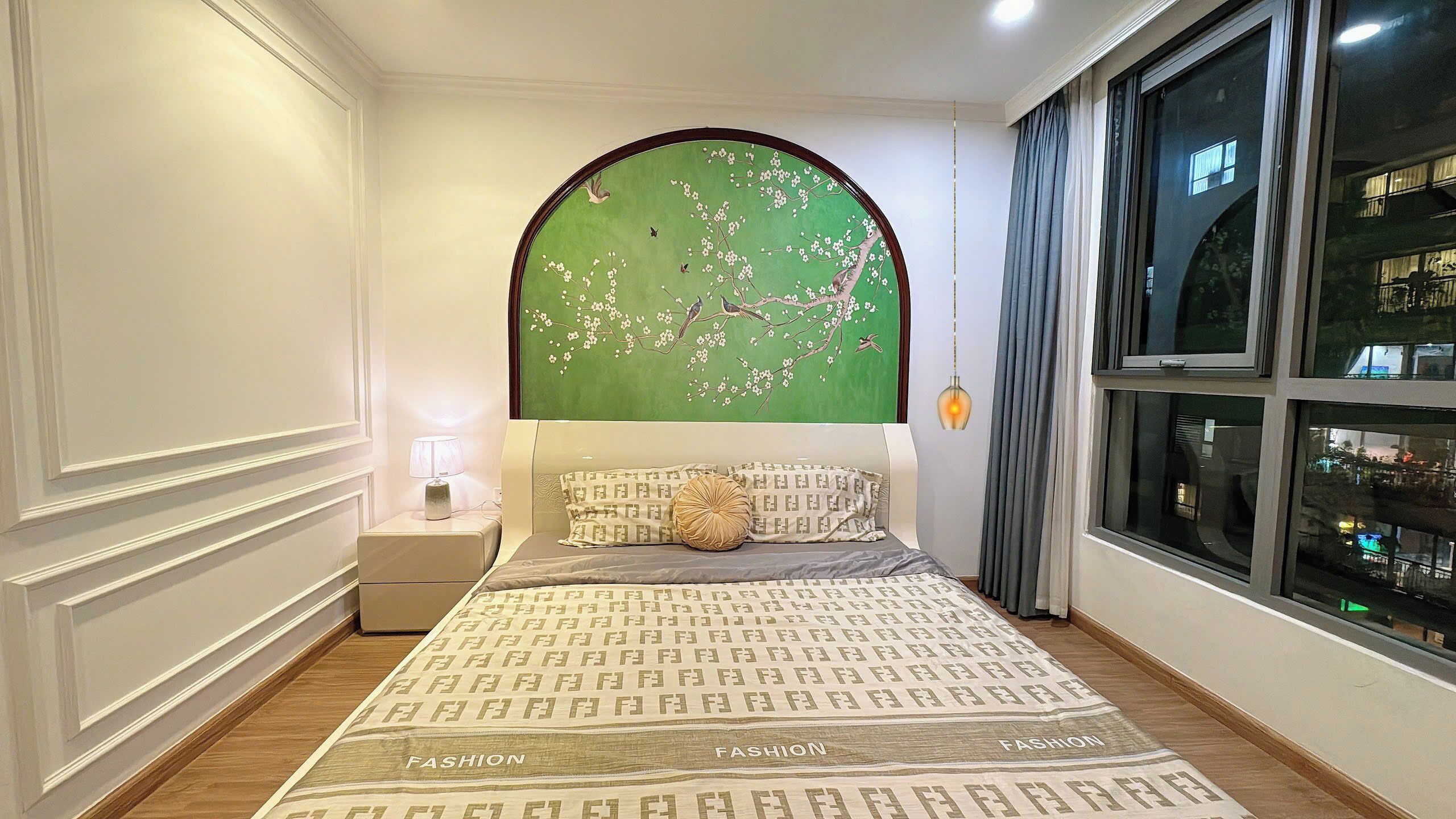 Un dormitorio con una cama grande, una mesita de noche, una lámpara, un muro verde con diseño floral y una ventana con cortinas.