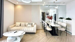 A modern living room with a sofa, coffee table, and dining area with chairs and a table. - Dani Apartment - Times City - Ha Noi (Hanoi)