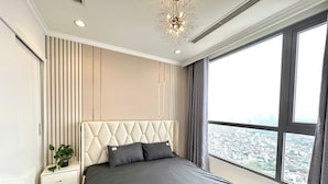 A modern bedroom with a large bed, a nightstand, a potted plant, and a city view through the window. - Dani Apartment - Times City - Ha Noi (Hanoi)