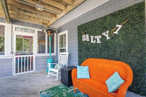 Eine überdachte Veranda mit einem 'WELCOME'-Schild, ein orangefarbenes Korbsitzsofa mit blauen Kissen, ein weißer Schaukelstuhl und eine grüne Wand mit den Buchstaben 'SALTY 7'.