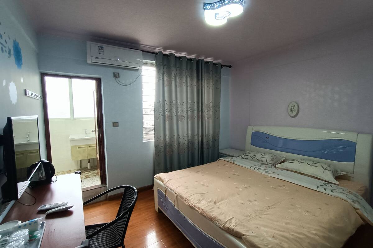 A bedroom with a bed, a desk with a chair, a mirror, a window with curtains, and an air conditioning unit.