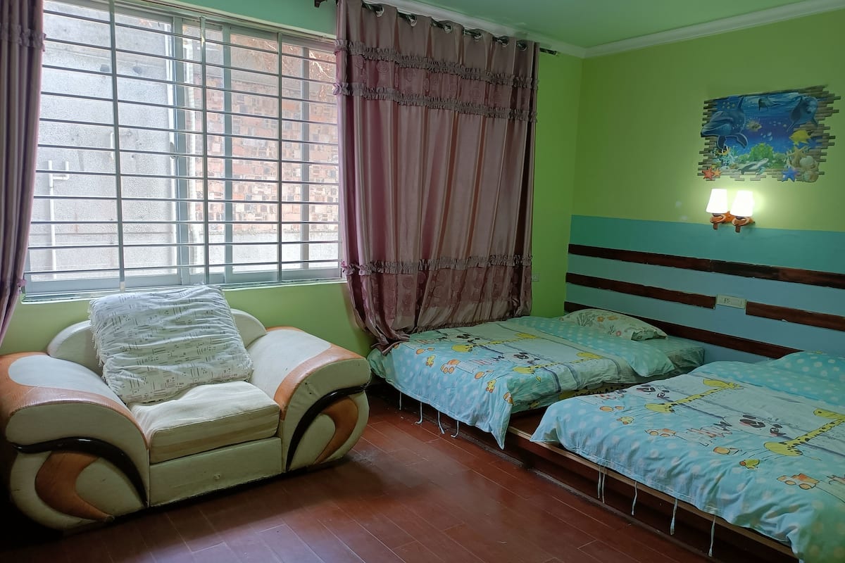 A room with a sofa, two beds, a window with curtains, and a wall painting.