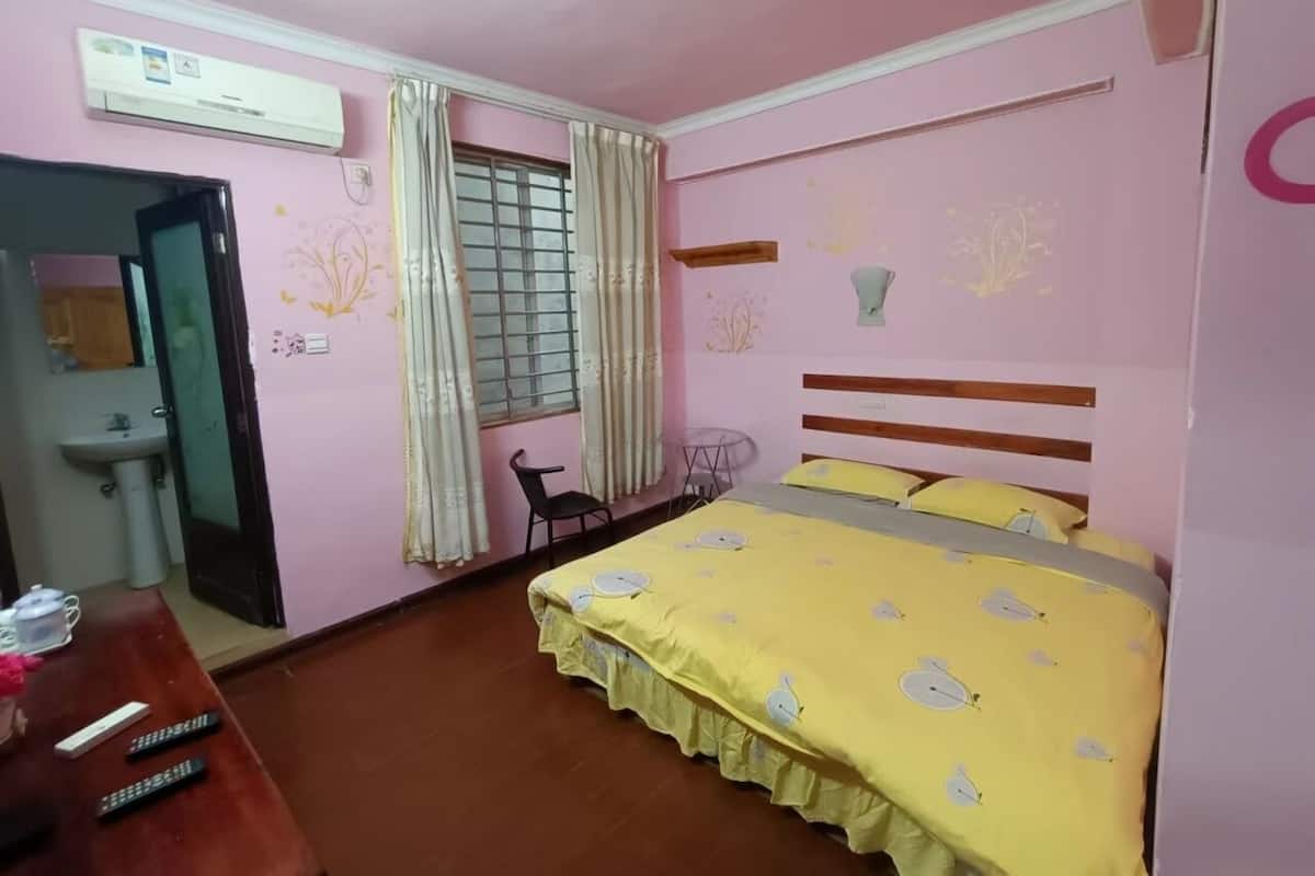 A bedroom with a bed, a desk, a chair, a window with curtains, a wall-mounted air conditioner, and a small table.
