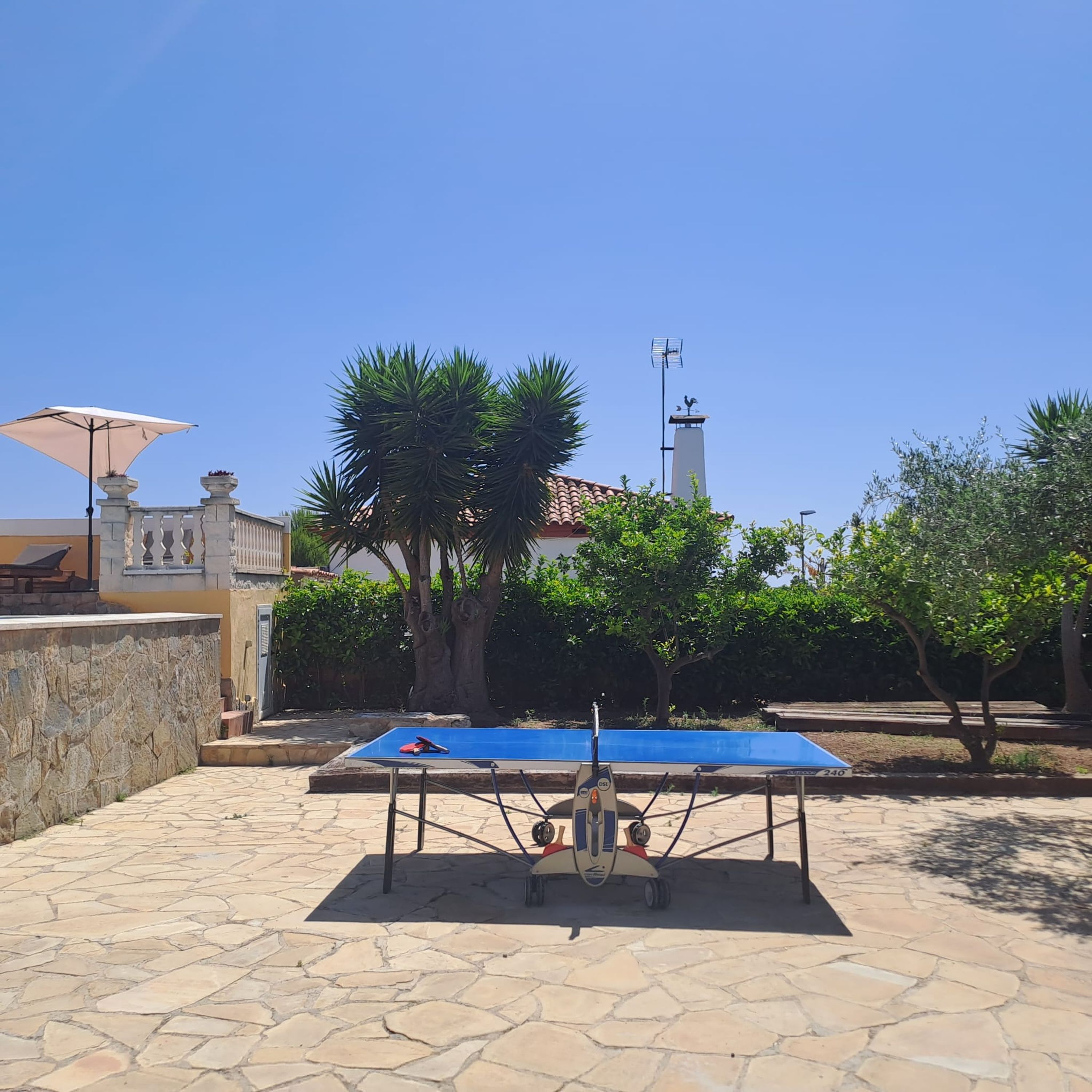 A ping pong table in an outdoor area with stone flooring, surrounded by trees and a building with a tiled roof.