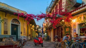 Una calle estrecha flanqueada por edificios, con una scooter roja estacionada en el centro y personas sentadas en mesas al aire libre.