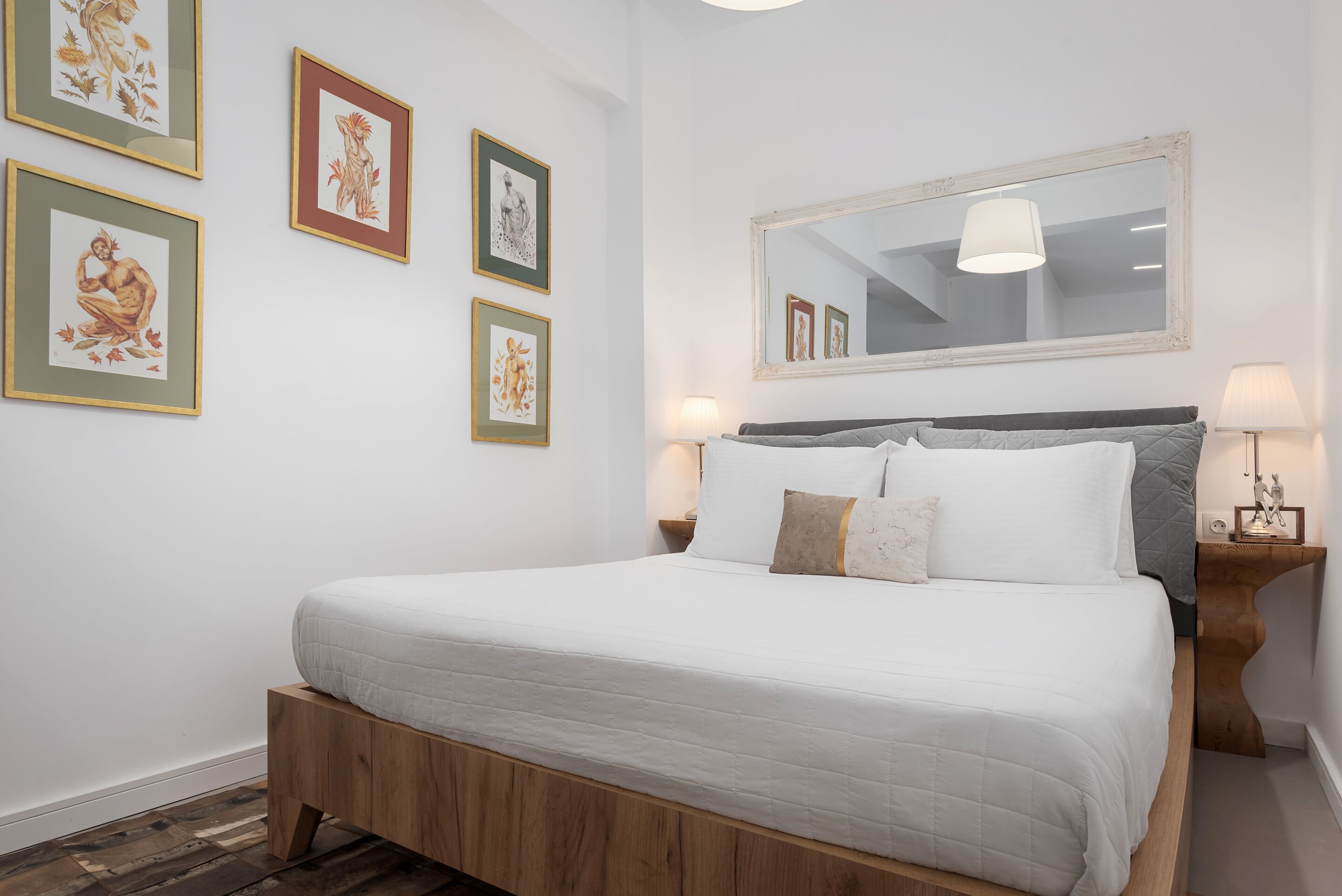 A neatly made bed with a wooden headboard, a large mirror above it, and four framed artworks on the wall.