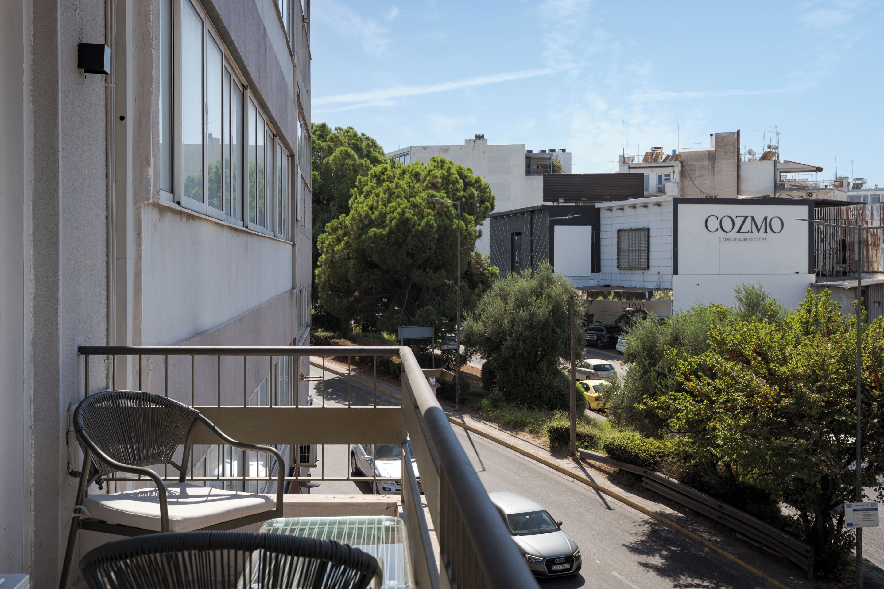 Un balcon avec deux chaises, une vue sur une rue où sont garées des voitures, et un bâtiment portant l’enseigne « COZMO ».