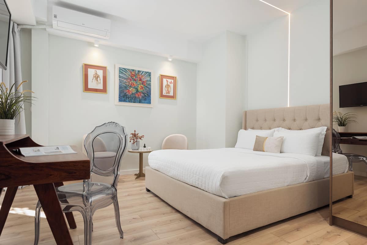 A modern bedroom with a large bed, a desk, and a chair. There are two framed artworks on the wall and a potted plant on the desk.