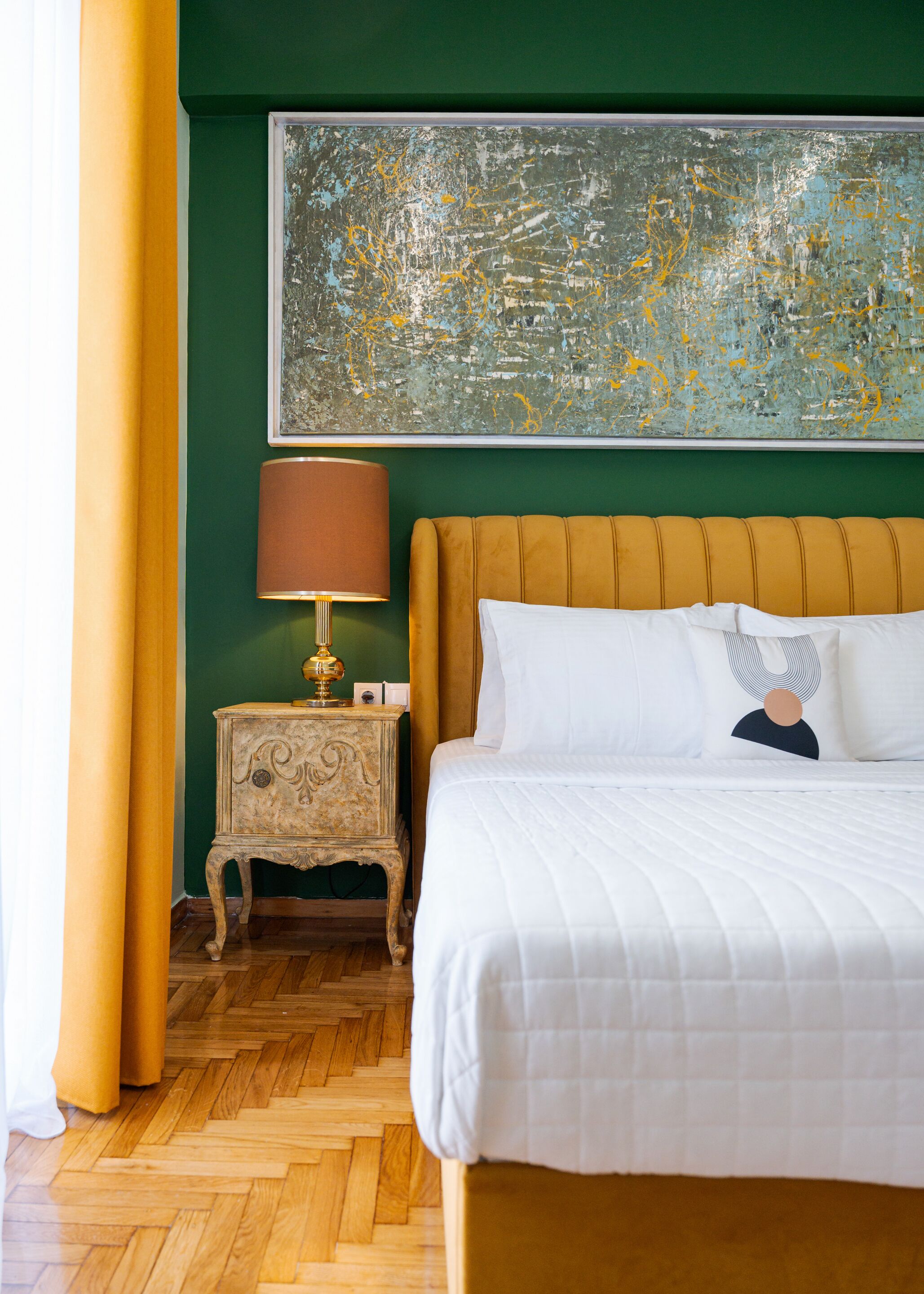 A hotel room with a large bed, a nightstand with a lamp, a green wall, and a large abstract painting.