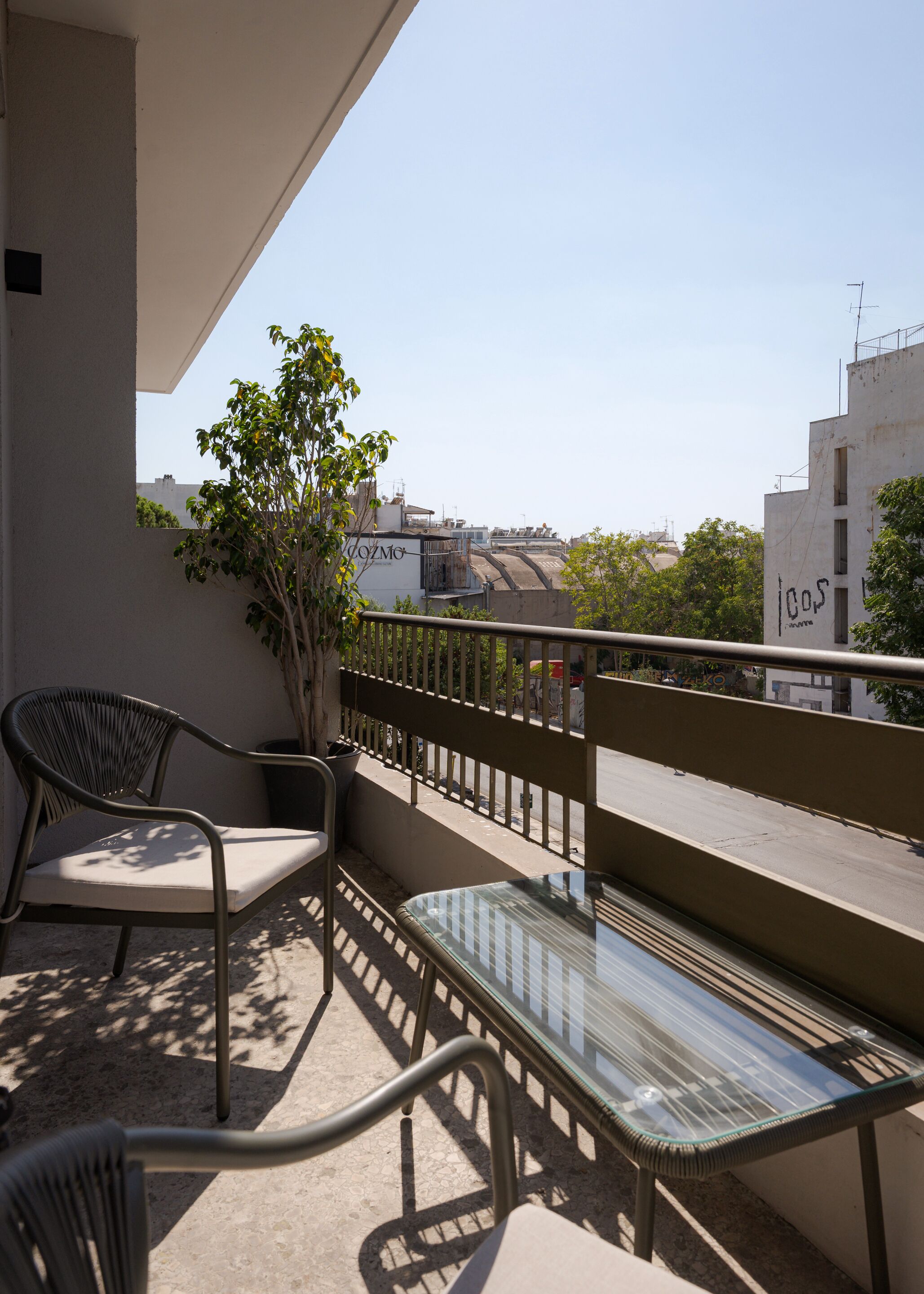 Un balcón con una mesa de vidrio y dos sillas, una planta en maceta y vistas a edificios en la distancia.