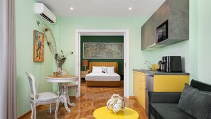 A hotel room with a bed, a dining area with a table and chairs, a sofa, a kitchen area with a microwave, and a large painting on the wall. - Olympian Zeus Residences (Athens)