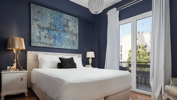 A bedroom with a large bed, two bedside tables, a chandelier, and a view of the outside balcony. - Olympian Zeus Residences (Athens)