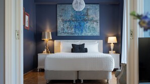 A bedroom with a large bed, two bedside tables with lamps, a chandelier, and a blue abstract painting on the wall. - Olympian Zeus Residences (Athens)