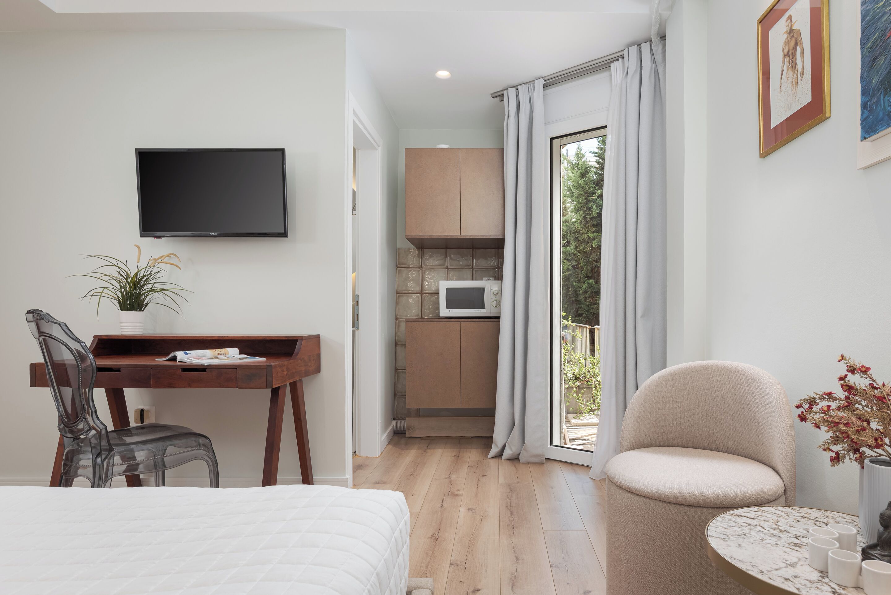 A modern hotel room with a bed, a desk with a chair, a television, a kitchenette, and a balcony with curtains.