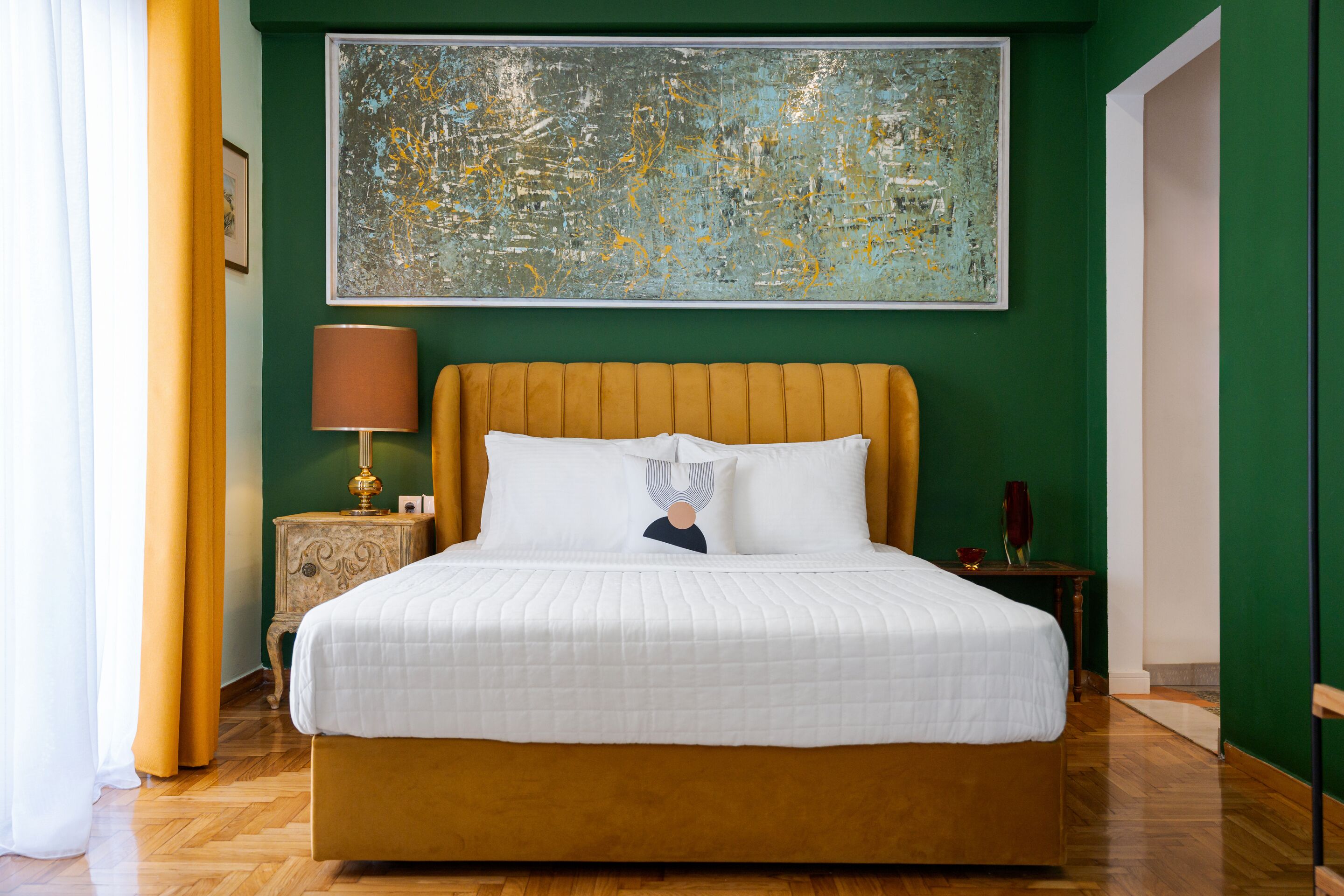 A neatly made bed with a white duvet and pillows, a wooden headboard, and a large abstract painting above it.