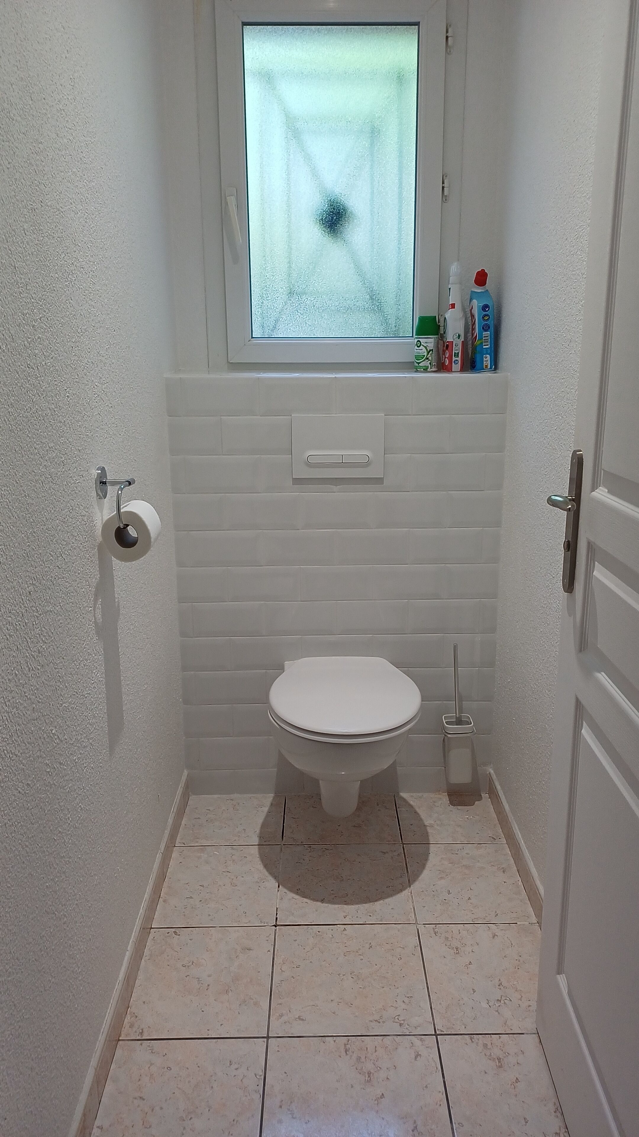 A small bathroom with a toilet, a window, and a shelf with toiletries.