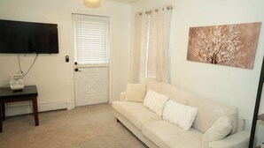 Una sala de estar con un sofá color beige, un televisor de pantalla plana montado en la pared, una puerta con ventanal y un cuadro de un árbol.