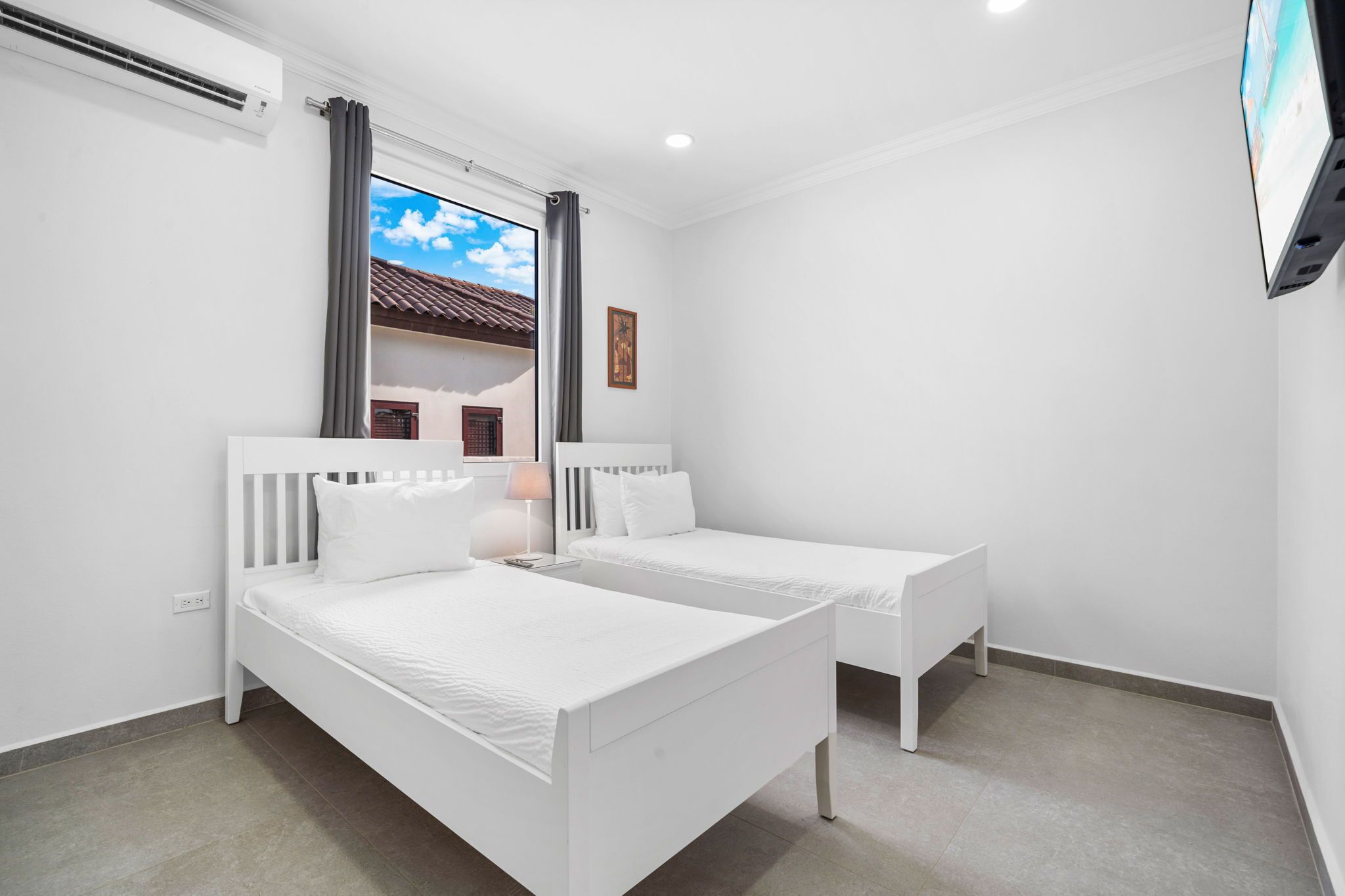 Quarto com duas camas brancas, uma janela com cortinas, uma TV na parede e uma pequena mesa de cabeceira com abajur.