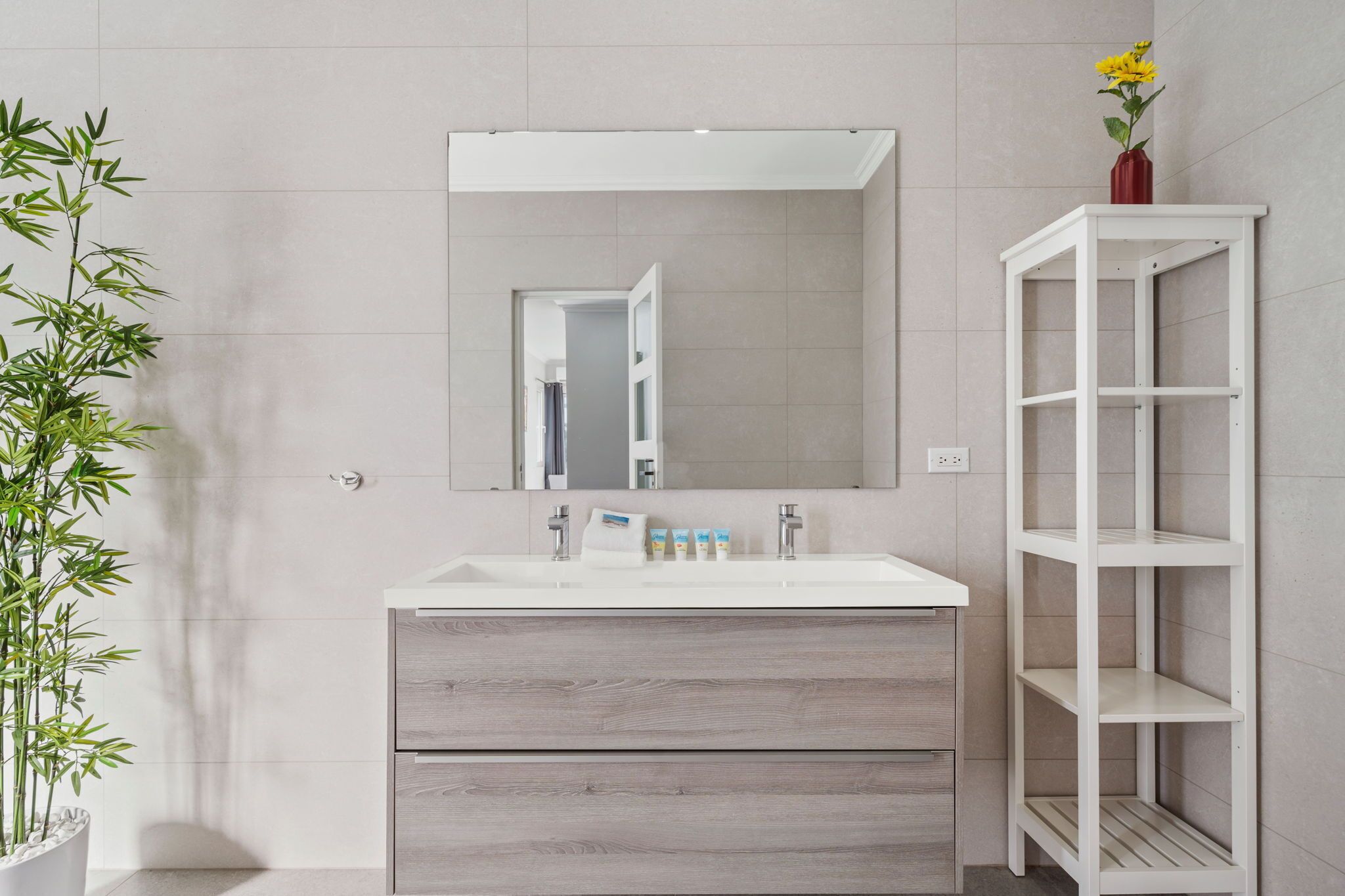 A bathroom with a white sink, mirror, and shelves. There is a bamboo plant and a vase with yellow flowers.