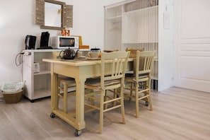 Cocina pequeña equipada con microondas, cafetera y tostadora. Hay una mesa con sillas, un cesto y una silla de mimbre.