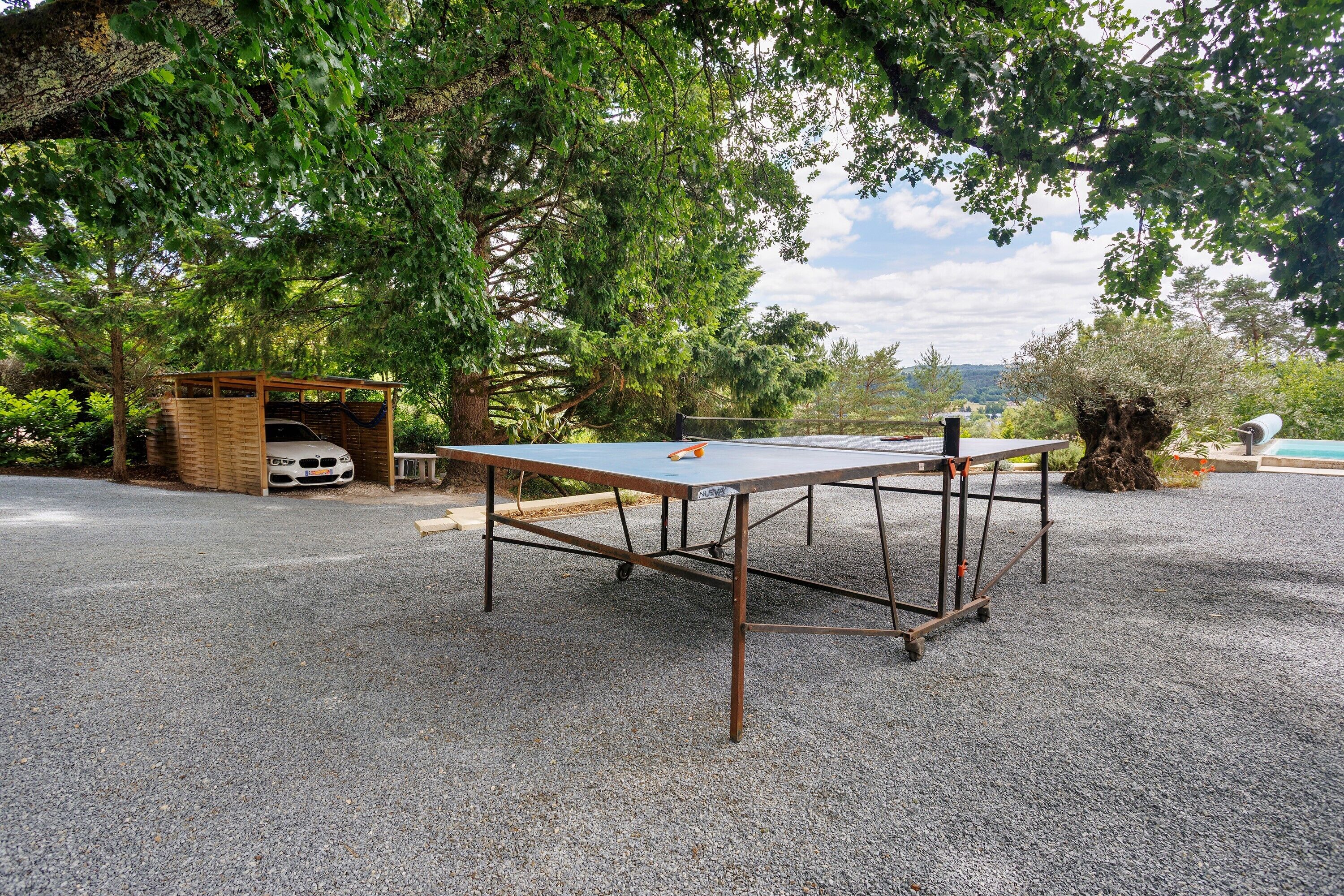 Una mesa de ping pong montada al aire libre sobre un terreno de grava, rodeada de árboles y una estructura de madera.