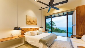 A modern bedroom with a large bed, a ceiling fan, a view of the ocean, and a wooden headboard.