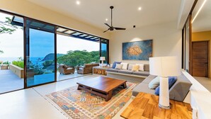 A spacious living room with a large sliding glass door leading to a balcony, a ceiling fan, and a modern sofa set.