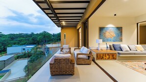 A modern living room with wicker furniture, a sofa, and a coffee table. There is a painting on the wall and a view of greenery outside.