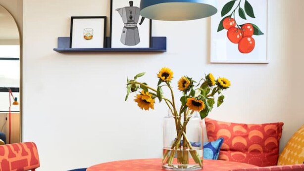 A vibrant room with a round red table, matching red chairs, and a blue hanging lamp. Sunflowers are in a clear vase on the table.