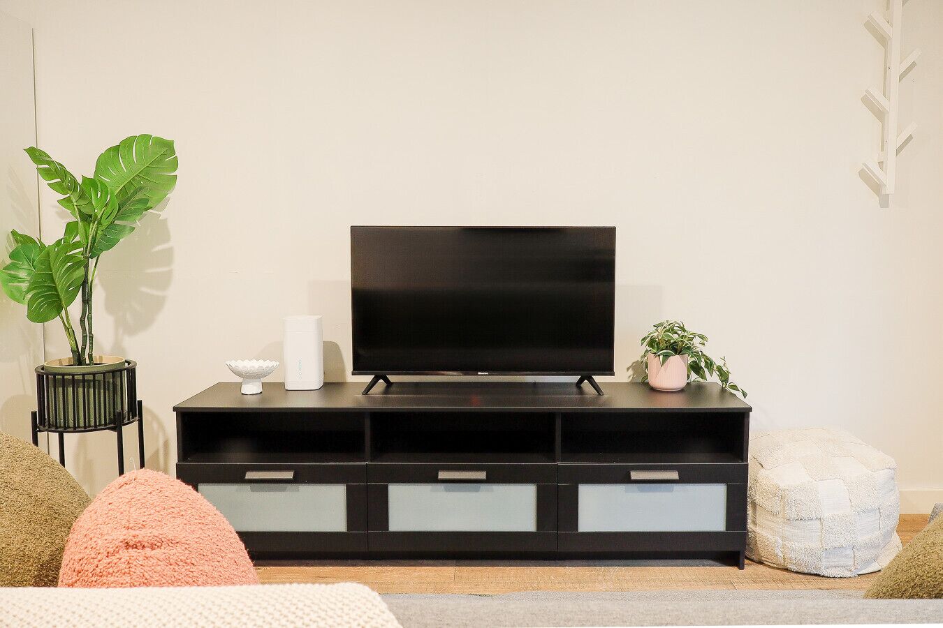 A modern living room with a flat-screen TV on a black entertainment unit, a potted plant, and a small white stool.