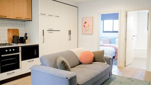 Une cuisine moderne avec des meubles blancs, un canapé gris agrémenté de coussins, et une vue sur une chambre à coucher avec un lit et une fenêtre.