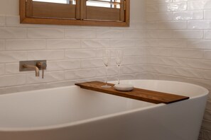 A modern bathroom with a freestanding bathtub, a wooden shelf with two glasses, and a wooden-framed window above. - Nancy’s of Trentham | Stylish 3BD Walk to Town (Trentham)