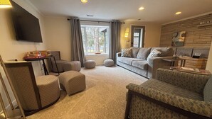A cozy living room with a sofa, ottomans, a coffee table, and a flat-screen TV mounted on the wall. - Stadium Stroll | Walk to IU Football | Sleeps 14 (Bloomington)
