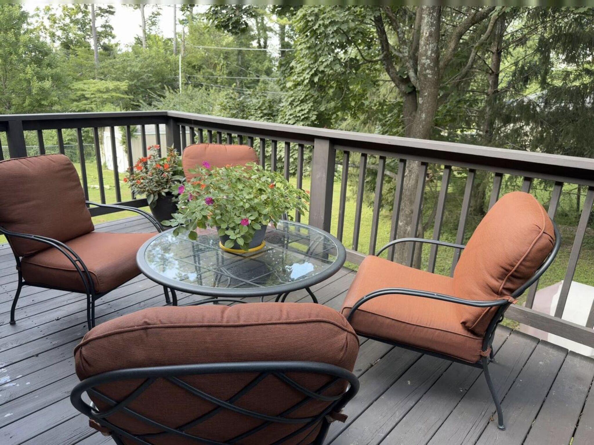 A deck with two brown cushioned chairs and a glass table, surrounded by a wooden railing and greenery.