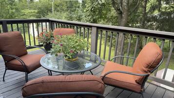 A deck with two brown cushioned chairs and a glass table, surrounded by a wooden railing and greenery.