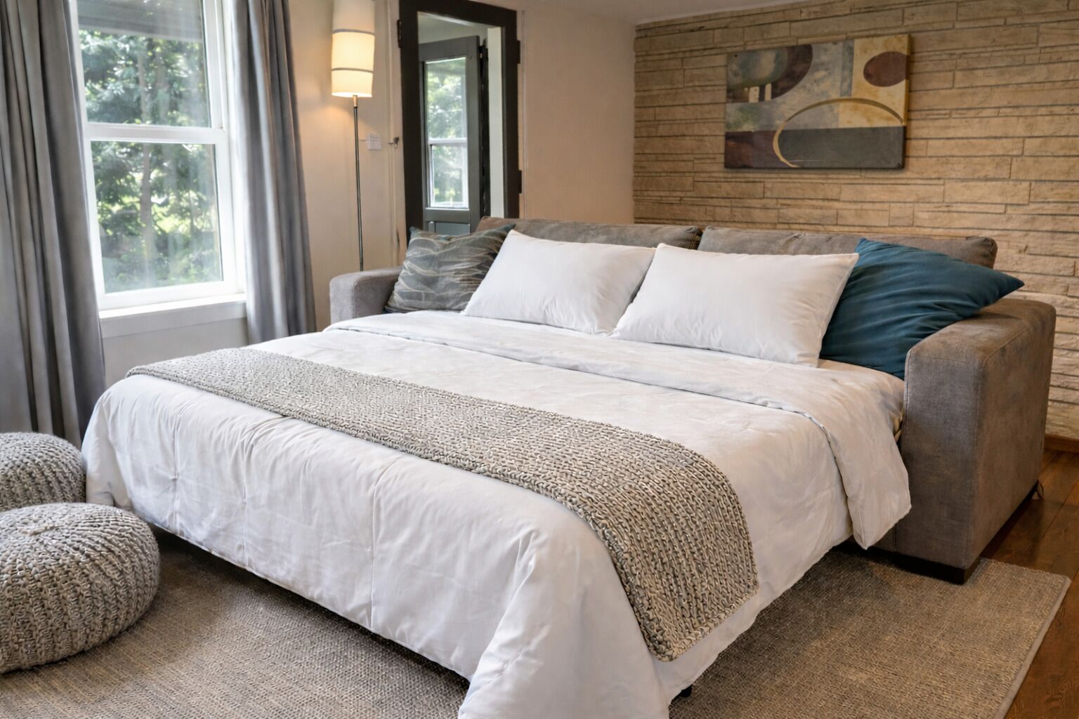 A bedroom with a large bed, a grey sofa, a woven throw blanket, and a stone wall with a painting.