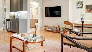 Uma sala de estar moderna com uma mesa de centro de vidro, um sofá, uma mesa de jantar com cadeiras, uma televisão fixada na parede e uma cozinha com armários e eletrodomésticos.