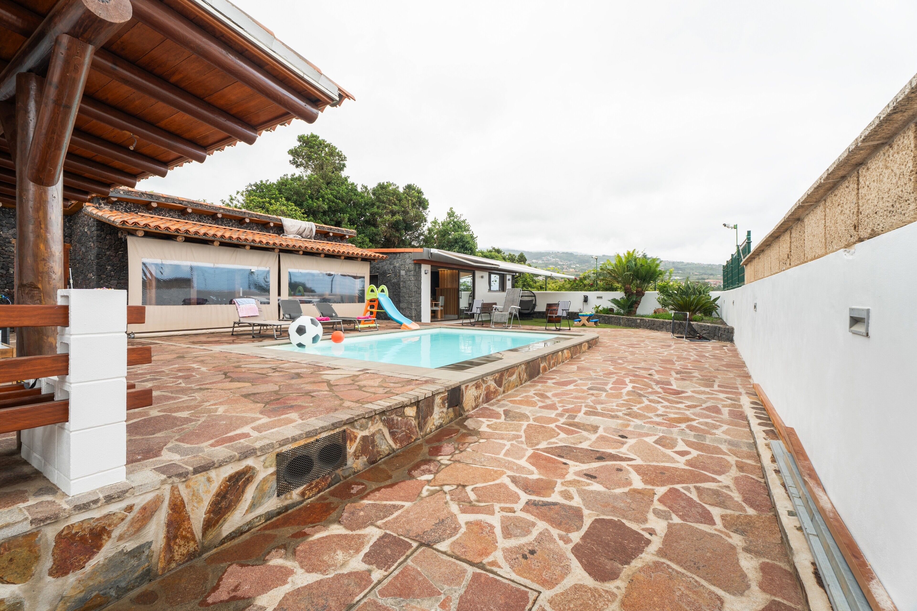 Un patio de piedra que conduce a una piscina con tobogán, un pérgola de madera y vistas del paisaje circundante.