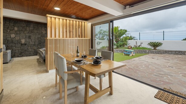 A modern dining area with a wooden table and chairs, a stone wall, and a view of a garden. - 'Vista Teide' Vacation Apartment with Sea View, Wi-Fi, and Air Conditioning (Tacoronte)