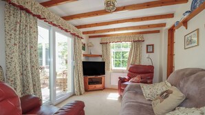 Una sala de estar acogedora con un sofá de cuero rojo, un sillón tapizado y un televisor sobre un mueble de madera. Amplias ventanas con cortinas florales dejan entrar abundante luz natural.