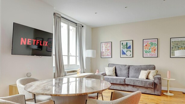Una sala de estar moderna con una televisión de pantalla plana, una mesa redonda de vidrio, un sofá gris y obras de arte enmarcadas en las paredes.