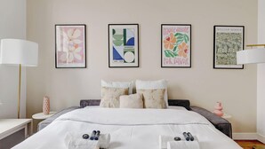 Habitación de hotel con cama, almohadas, mesita de noche, lámpara y cuatro cuadros enmarcados en la pared.
