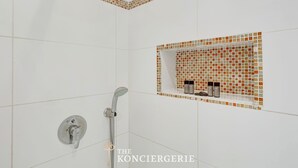 Un baño moderno con azulejos blancos, ducha con cabezal de ducha manual y estante con artículos de aseo.