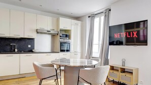 Una cocina moderna con armarios blancos, horno empotrado y microondas. Hay una mesa de comedor con sillas y un televisor de pantalla plana montado en la pared mostrando 'NETFLIX'.