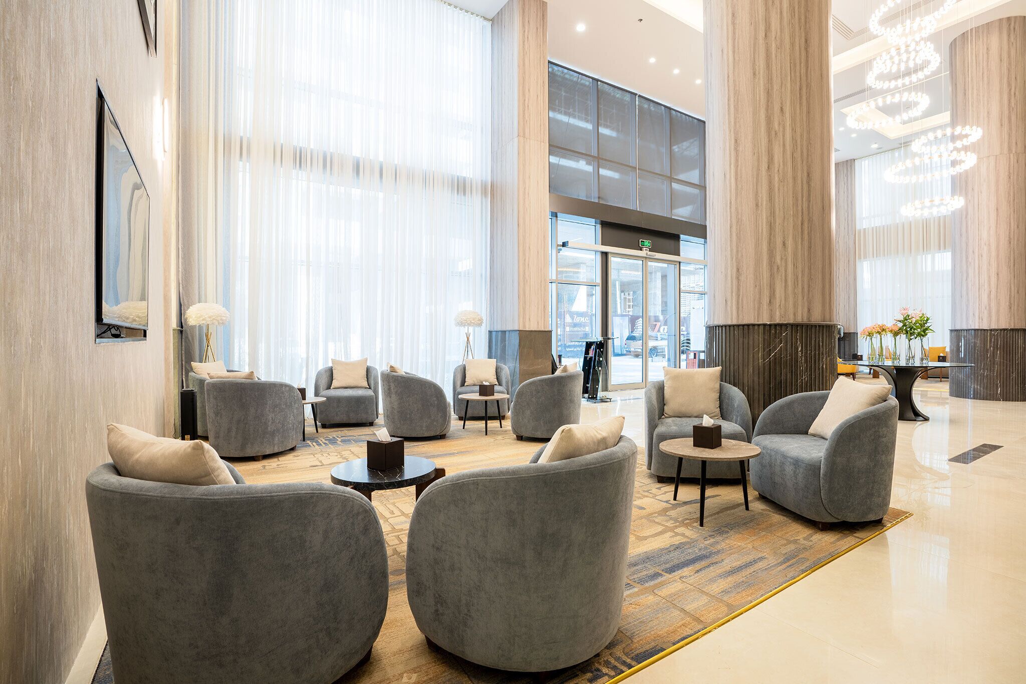 A spacious lobby with grey upholstered chairs, a central coffee table, and a large window with sheer curtains.